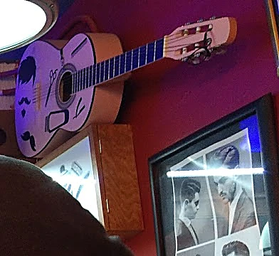 Guitarra blanca pintada con bigotes y tijeras, colgada en la pared roja junto a un cuadro de cortes clásicos