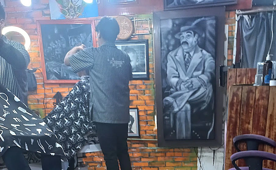 Interior de la barbería: pared de ladrillo naranja, cuadro en blanco y negro de un gánster de traje, un barbero de espaldas con camisola de rayas y clientes cubiertos con la capa negra de la casa