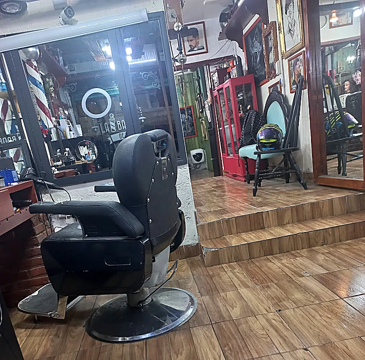 Sillón de barbero negro sobre el piso de duela, escalones, vitrina roja de época y una silla turquesa al fondo; en el vidrio, el logo esmerilado con el poste